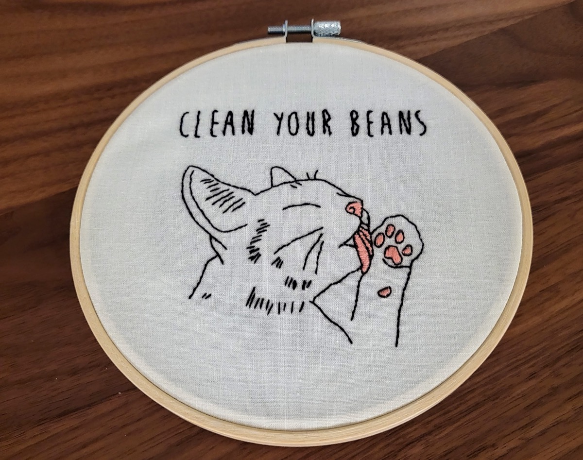 Backstitch and satin stitch embroidery. A cat licking its paw, with the caption &lsquo;Clean Your Beans&rsquo;.